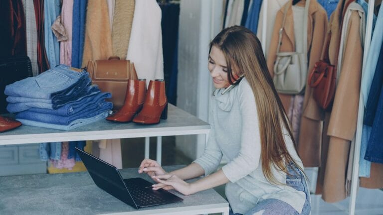 Shop owner managing digital marketing for small business on a laptop and phone in her clothing store